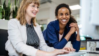 two women sitting at a table with a laptop