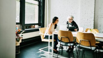 two women sitting at a table with a laptop